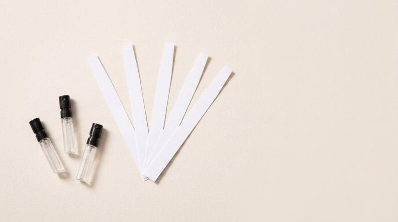 A fan of white paper blotter strips beside three small 2ml perfume sample vials on warm off-white paper