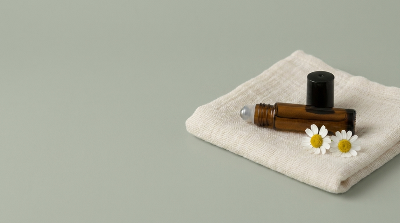 A 20.O2 amber glass roll-on vial resting on folded cotton gauze with chamomile flowers on a soft sage background